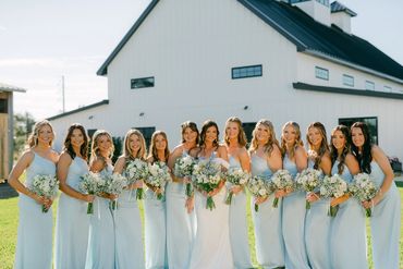 Bride with bridesmaids in light blue dresses holding bouquets outdoors.