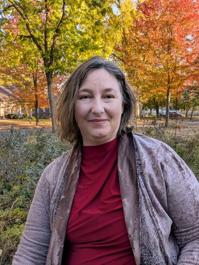 Woman smiling outdoors with autumn foliage in the background.