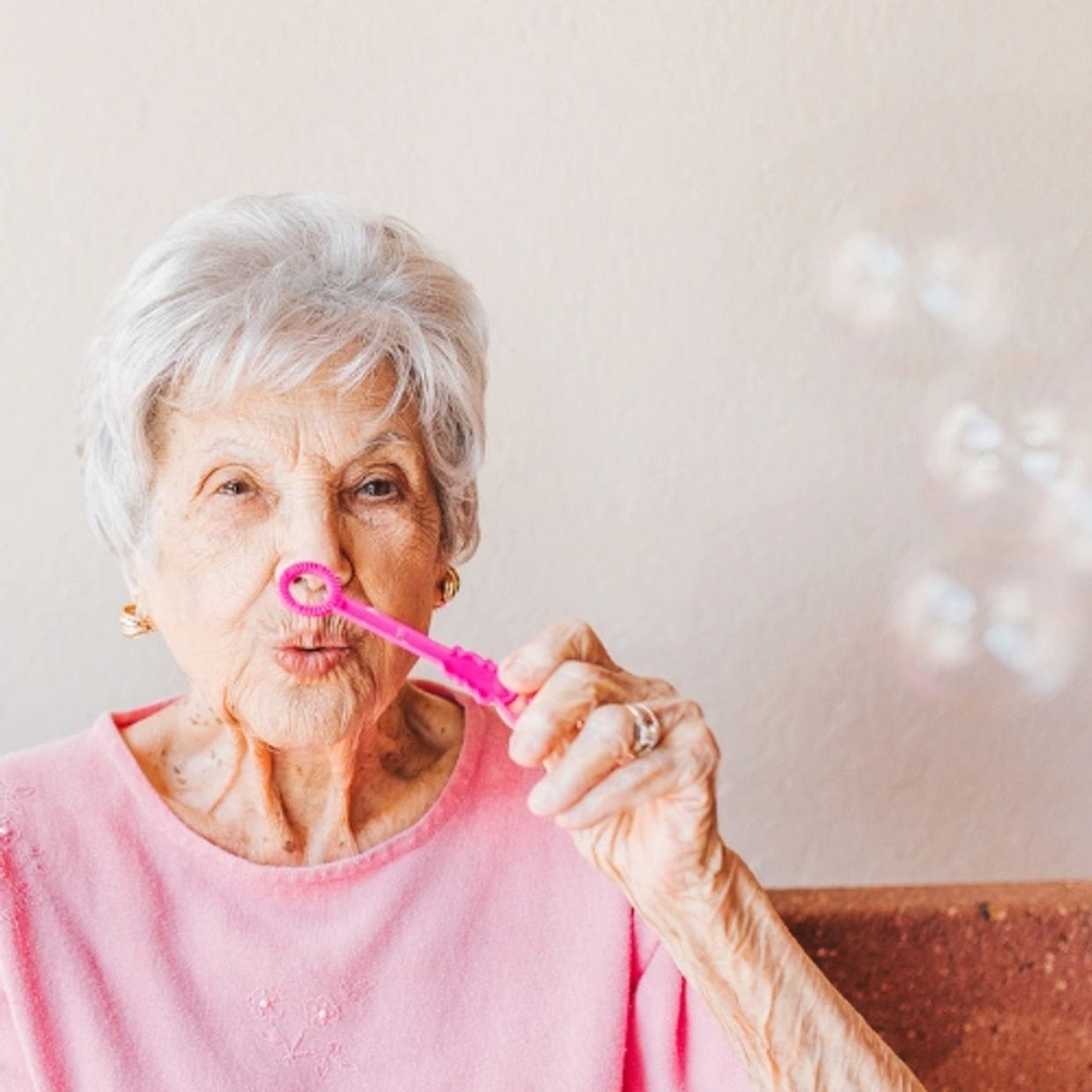 Elderly woman blowing bubbles with a pink wand indoors.