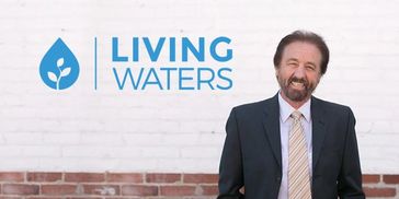 Man in suit smiling beside Living Waters logo on a brick wall.