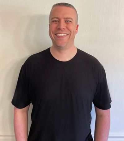 Smiling man with short hair wearing a black t-shirt against a light wall.