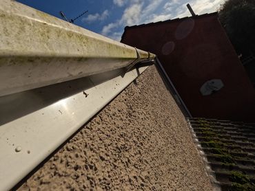 Close-up of a roof edge with gutter and textured wall under a partly cloudy sky.
