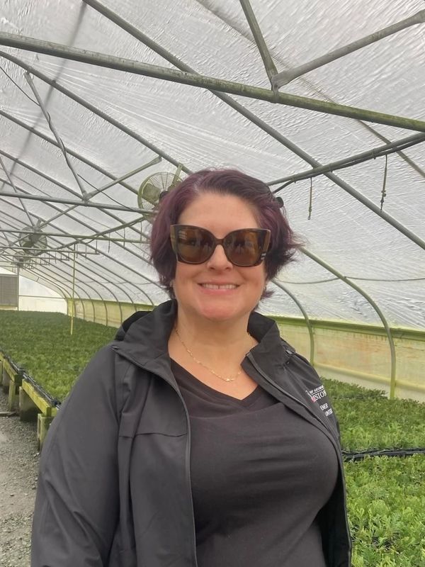 Smiling woman with sunglasses inside a greenhouse filled with plants.