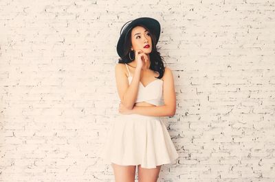 Woman in a white dress and black hat posing against a white brick wall.