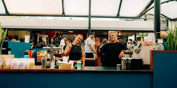 Two baristas smiling and waving behind a coffee counter in a busy cafe.