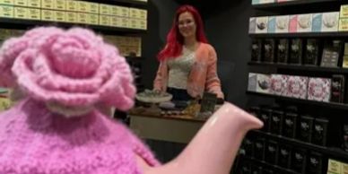 Pink teapot with crocheted cozy and a woman in the background.