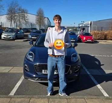 Young man standing in front of a dark blue car holding a smiley face emoji.
