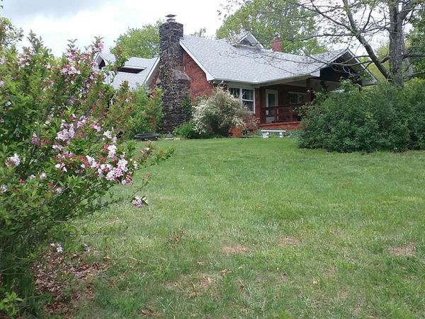 Cozy brick house with a stone chimney surrounded by greenery and blooming flowers.