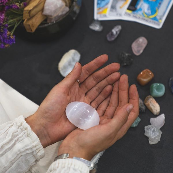 Hands holding a smooth white crystal with other stones and tarot cards nearby.