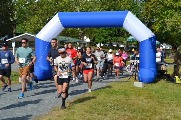 Runners start a race under a blue and white inflatable arch on a sunny day.
