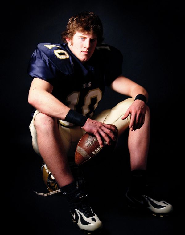 Young football player in navy and gold uniform holding a football, seated against a dark background.