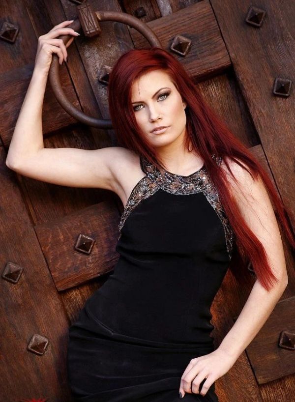 Woman with long red hair in a black dress posing by a rustic wooden door.