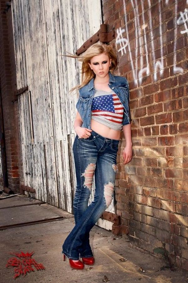 Young woman in ripped jeans and American flag top leans on brick wall.
