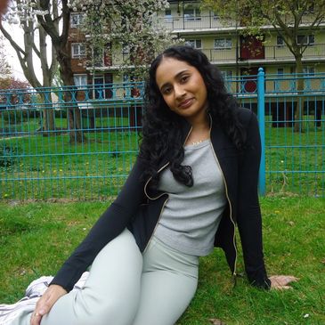 A woman with long curly hair sitting on grass in a park, smiling gently.