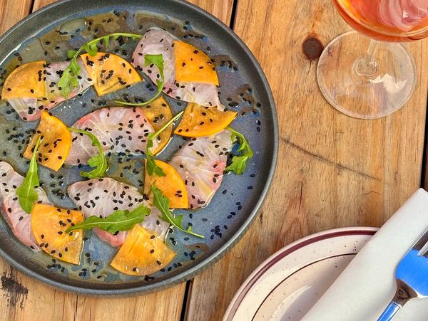Artistic plate of sashimi with orange slices, greens, and black sesame seeds on wood table.