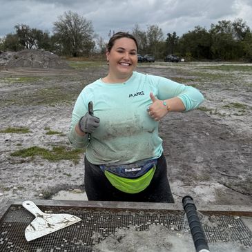 Smiling woman giving thumbs up while working outdoors in muddy conditions.