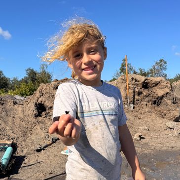 Smiling child holding a small object in a muddy outdoor area with shovels in the background.