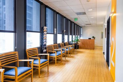 A Waiting room with blue and wood chairs against a wall of windows. 