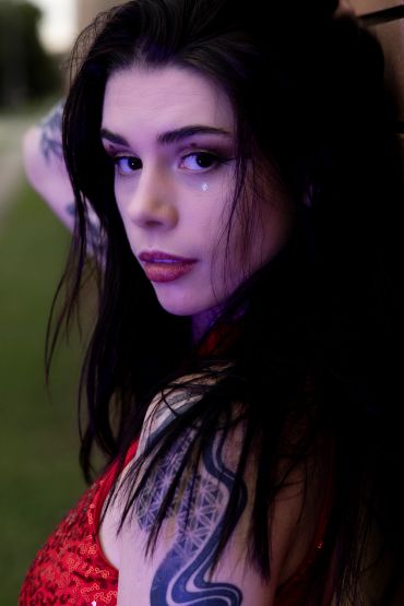 Young woman with dark hair and tattoos, wearing a red sequined top.