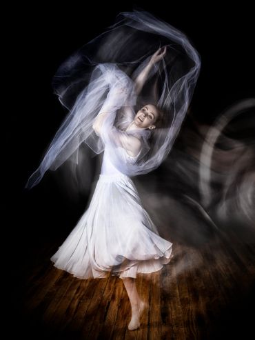 A dancer in white gracefully twirls with flowing fabric against a dark background.