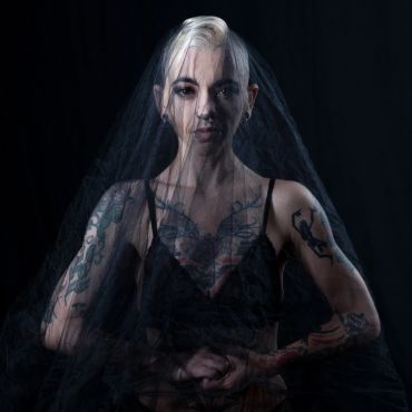 Tattooed woman draped in black veil against dark background.