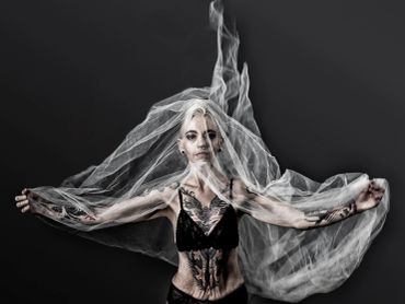A woman with tattoos poses under a flowing translucent veil against a dark background.