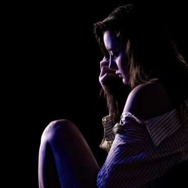 A contemplative woman sits alone in low light, deep in thought.