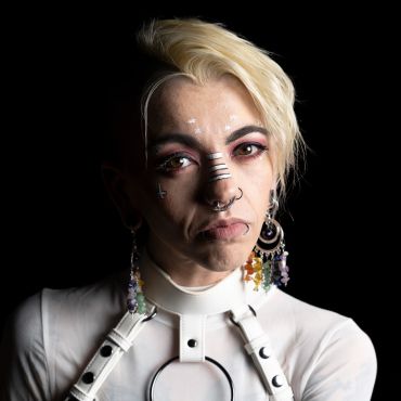 Person with blonde hair and unique facial piercings against black background.