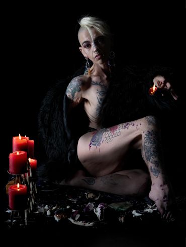 Tattooed person in dramatic lighting with red candles and dark fur.