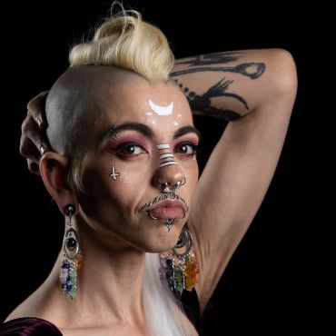 Person with a unique hairstyle, facial piercings, and artistic face paint posing against a black background.