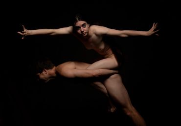 Two nude individuals performing an artistic pose against a black background.
