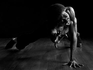 A muscular man striking a powerful dance pose on a wooden floor in dramatic lighting.