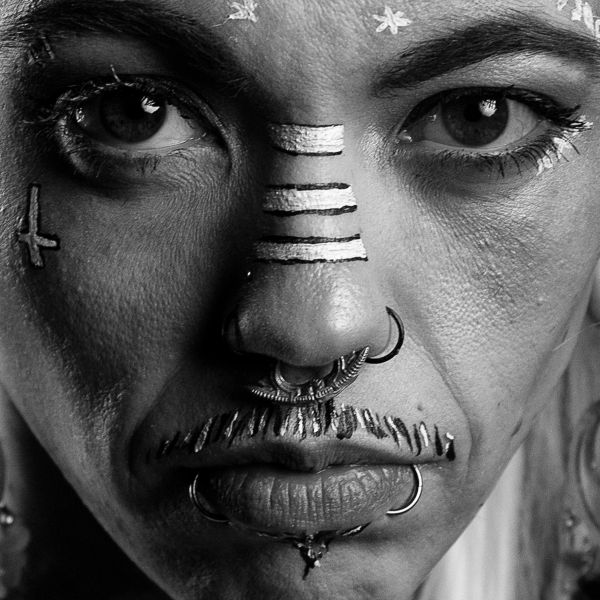 Close-up of a face with tribal-style paint and multiple facial piercings in black and white.
