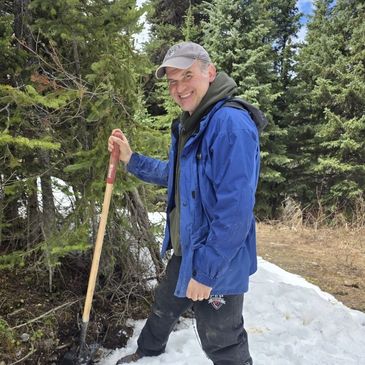 Man in blue jacket holding shovel in snowy forest.
