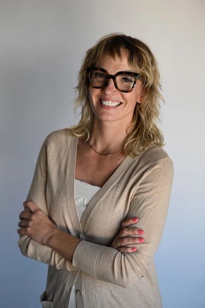 Smiling woman with glasses and a beige cardigan stands with arms crossed.