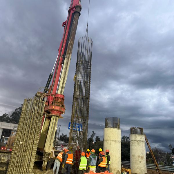 Construction workers hoisting steel reinforcement cage with crane at a cloudy site.