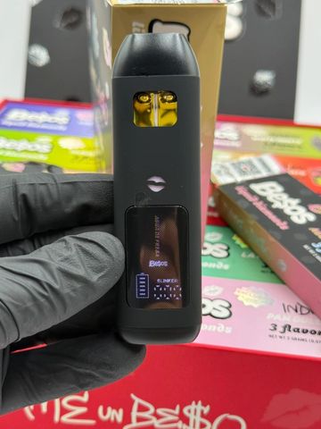 Hand holding a black vape device with colorful packaging in the background.