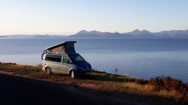 Campervan with raised roof parked by a lake and mountains.