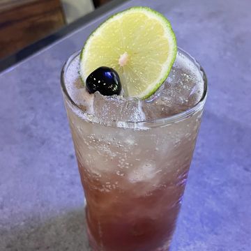 Tall glass with icy cocktail and garnished with lemon and Luxardo cherry.