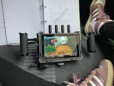 Remote control device with screen showing a camera feed, held between two hands with sneakers visible.