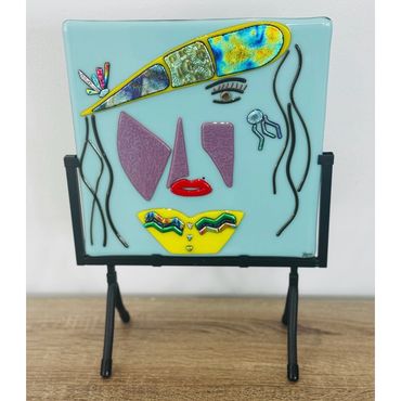 Abstract mosaic face art on a blue glass panel with a stand.
