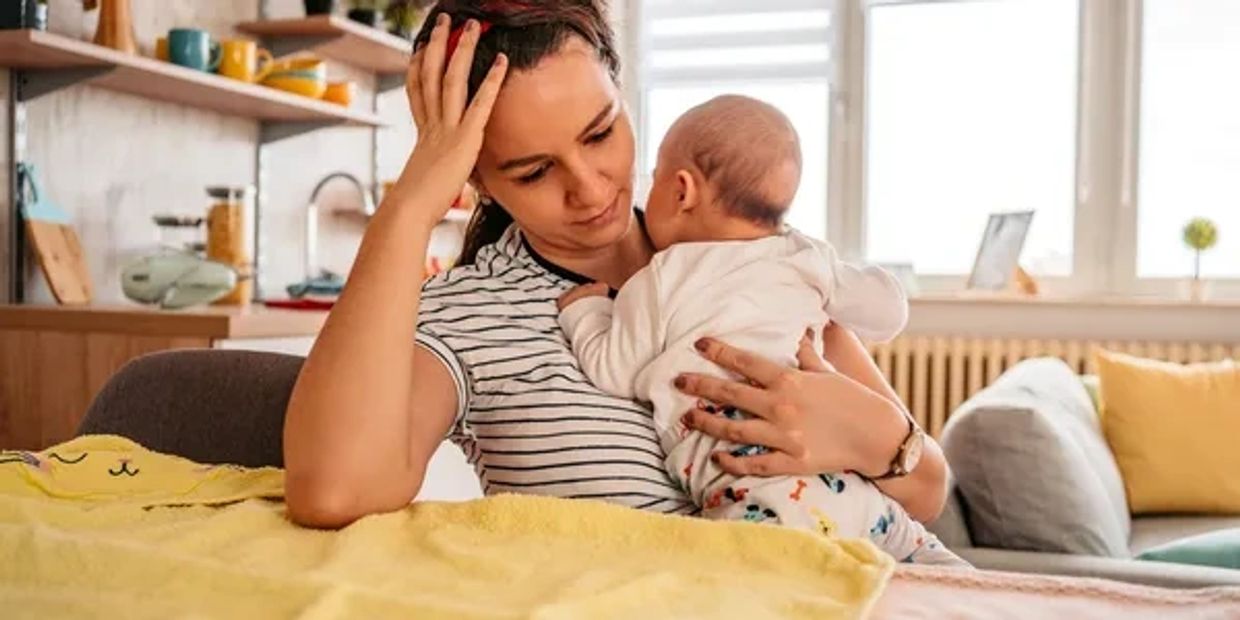 A tired mother holds her baby close while sitting in a cozy room.