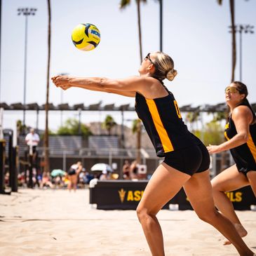 Female volleyball player in black and yellow uniform lunging to hit a ball on a sunny beach court.