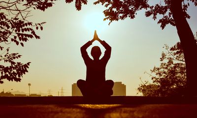 cross-legged woman with hands up in prayer pose with sun shinning out in nature next to a tree