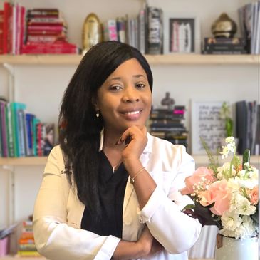 Smiling woman in white coat with flowers and books behind her.