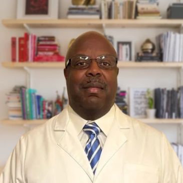 Man in white coat and glasses stands in front of a bookshelf.