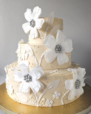 Elegant three-tier beige cake adorned with large white flowers and pearl-like decorations.
