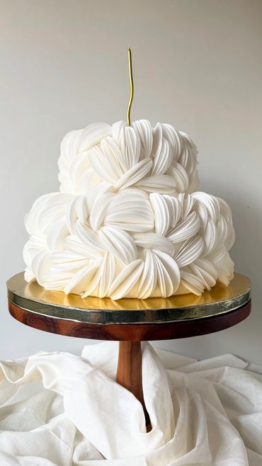 Elegant white tiered cake with intricate petal-like frosting on a wooden stand.