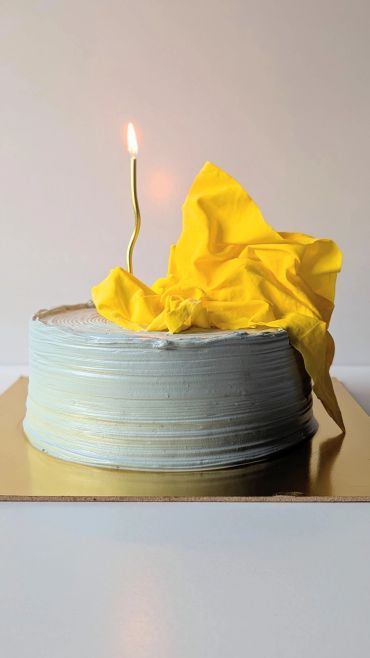 Minimalist cake with blue icing, a yellow cloth decoration, and a lit curved candle.