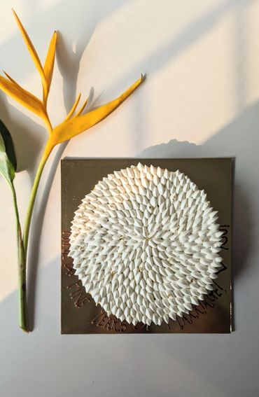A white, textured circular art piece beside a yellow bird of paradise flower on a white surface.